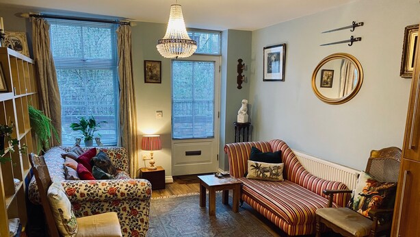 A cozy living room with a sofa, armchair, and a wooden coffee table. There is a chandelier hanging from the ceiling, and a large window with curtains. - Beautiful Town Centre Apartment With River Terrace (Macclesfield)