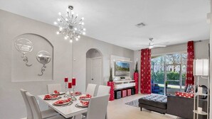 Un comedor moderno con una araña, una mesa de comedor para cuatro personas, y una sala de estar con un sofá y un televisor.