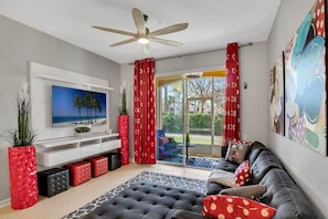 A modern living room with a flat-screen TV, a ceiling fan, a sofa with patterned cushions, and a large window with red curtains.