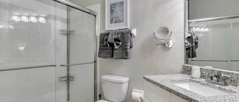 A bathroom with a glass-enclosed shower, a toilet, a sink with a granite countertop, and a mirror.