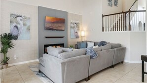 Ein Wohnzimmer mit grauem Sofa, Kamin und Wandbildern, darunter ein Meeresblick und zwei Vogelgemälde.