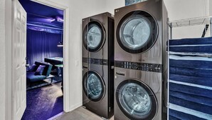 A laundry room with two stacked front-loading washing machines, a sign above indicating it's a laundry area, and a glimpse of a room with a pool table and blue curtains. - Luxurious home 2 miles from Disney - Windsor Hills Resort (Kissimmee)