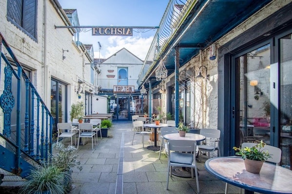 A narrow outdoor corridor with tables and chairs, a staircase, and a building with a sign reading "FUCHSIA".