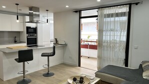 A modern kitchen with white cabinets, a black countertop, and stainless steel appliances. There is a dining area with two black bar stools and a wooden table with decorative items. A large sliding glass door leads to an outdoor balcony with plants. - 2 BED DUPLEX APARTMENT IN THE HEART OF LAS AMERICAS (Playa Delas Americas)