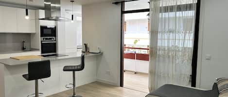 A modern kitchen with white cabinets, a black countertop, and stainless steel appliances. There is a dining area with two black bar stools and a wooden table with decorative items. A large sliding glass door leads to an outdoor balcony with plants.