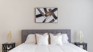 Une chambre à coucher moderne avec un grand lit, une tête de lit grise, du linge de lit blanc et une œuvre d’art abstraite accrochée au mur.