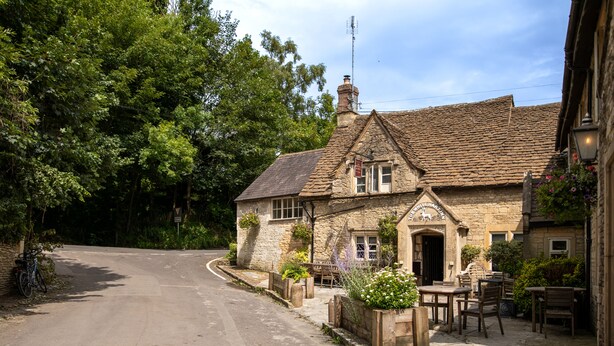 A stone building with a tiled roof, a wooden door, and a small porch with outdoor seating. - The White Hart (Chippenham)