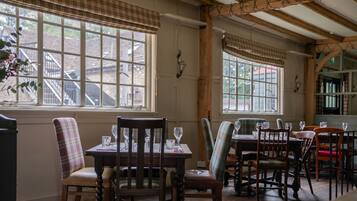 A dining area with wooden tables and chairs, large windows with curtains, and a view of the outside.