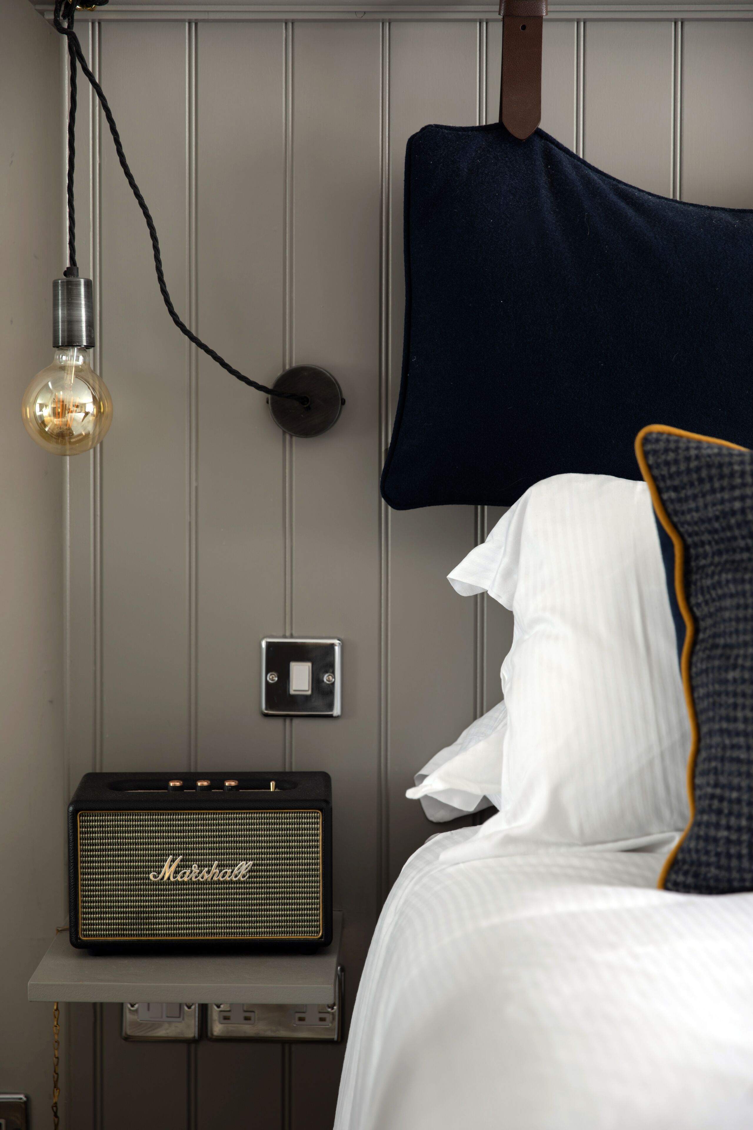 A bed with white bedding, a dark blue pillow, a Marshall speaker on a small table, and a light bulb hanging from the ceiling.