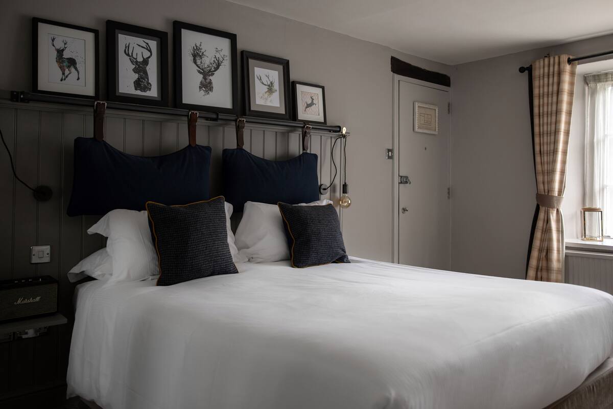 Una cama bien hecha con sábanas blancas y almohadas azul oscuro, una obra de arte con ciervo enmarcada sobre la cama y una cortina a cuadros en la ventana.