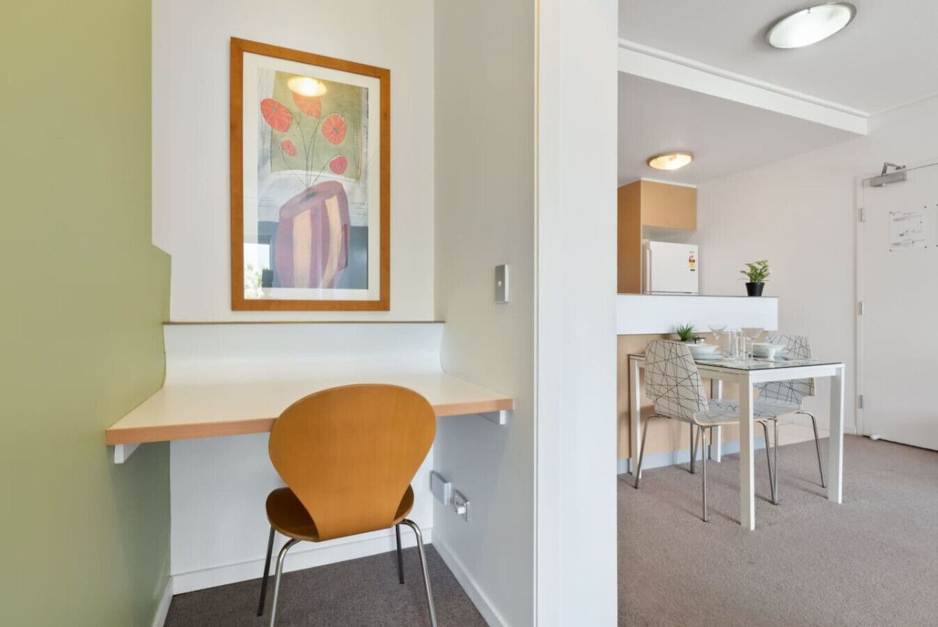 A modern room with a wooden desk, a single orange chair, a dining area with a glass table and chairs, and a framed artwork on the wall.