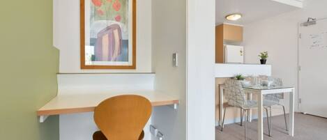 A modern room with a wooden desk, a single orange chair, a dining area with a glass table and chairs, and a framed artwork on the wall.