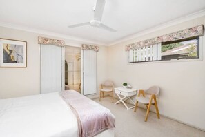 A bedroom with a bed, a desk, two chairs, a ceiling fan, and a window with a view.