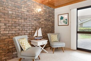 Una habitación con paredes de ladrillo, techo de madera, dos sillas grises con cojines estampados, una mesita con un modelo de velero y un cuadro en la pared.