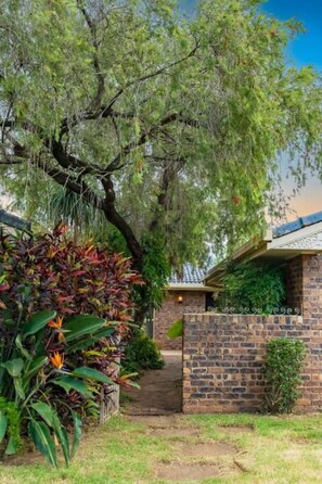 Una propiedad residencial con fachada de ladrillo, un acceso cubierto y una variedad de vegetación, incluyendo un árbol grande y plantas con flores.