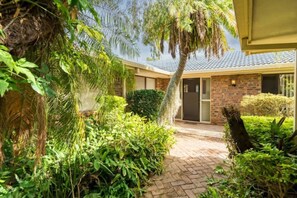 A residential building with a brick facade, a paved entrance, and a variety of greenery including a palm tree and shrubs.