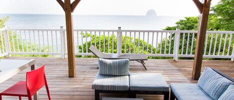 A covered outdoor seating area with a view of the sea, wooden deck, and white railings.