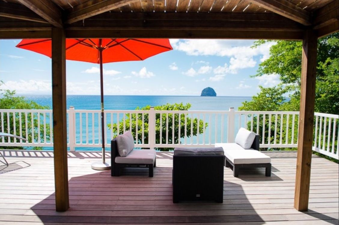 Une terrasse couverte avec vue sur l’océan, comprenant deux chaises longues et un parasol rouge.