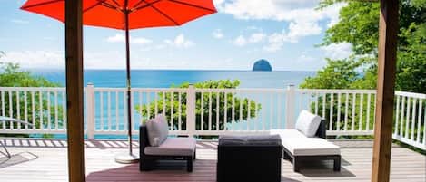 Une terrasse couverte avec vue sur l’océan, comprenant deux chaises longues et un parasol rouge.