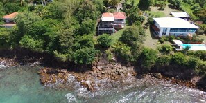 An aerial view of a coastal property with multiple buildings, lush greenery, and a rocky shoreline.