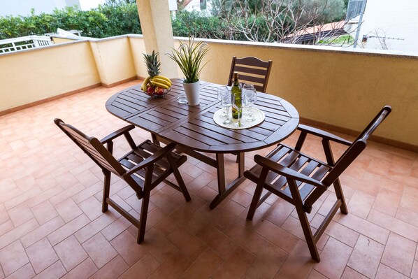 A round wooden table with four chairs, a potted plant, and a bottle of wine on a balcony with terracotta tiles.