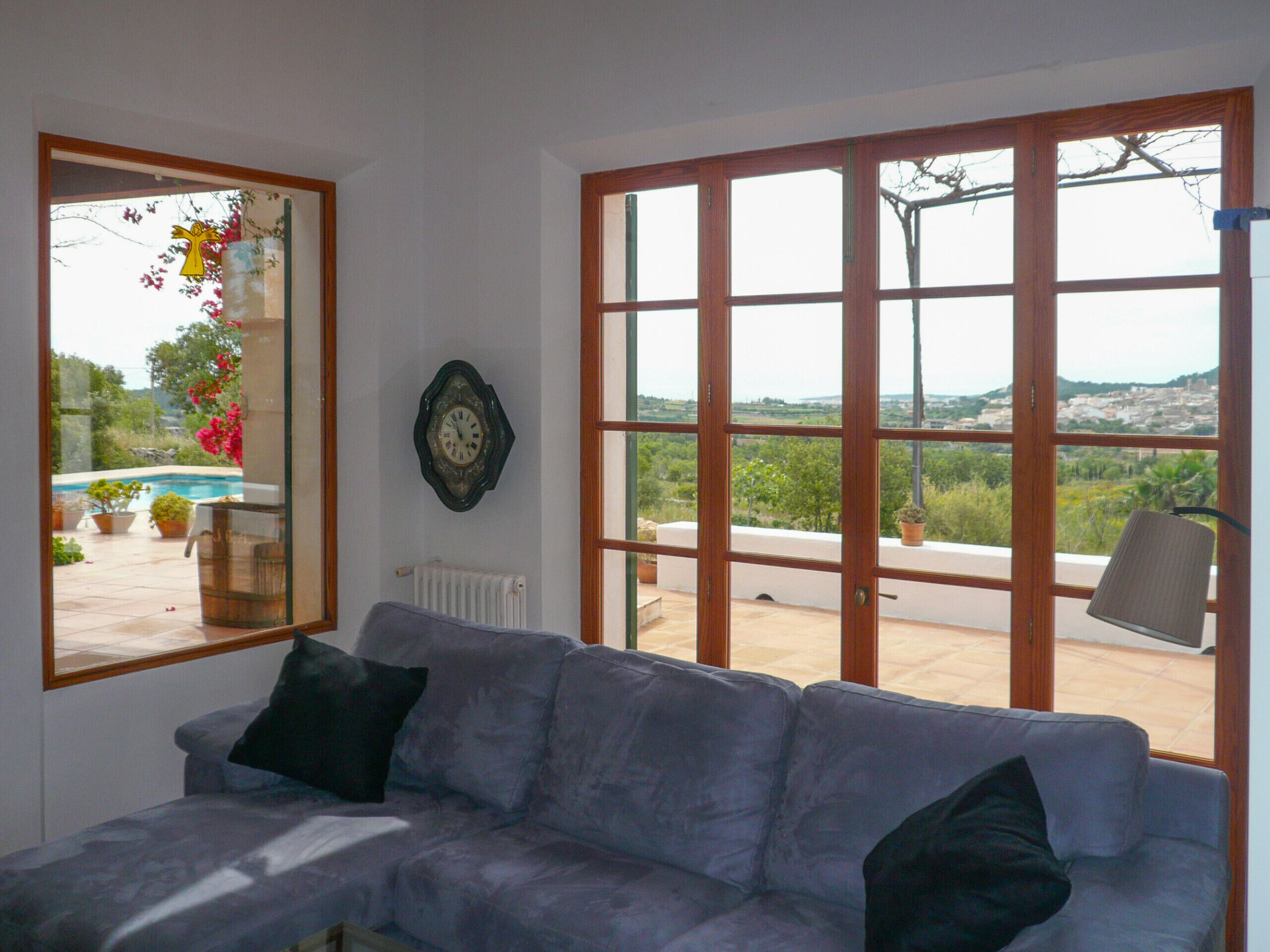 Une pièce avec un canapé gris, une fenêtre à cadre en bois et une vue sur une piscine et la verdure environnante.