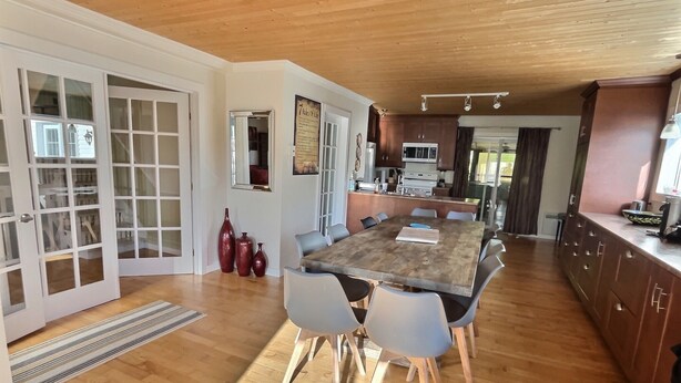 Un vaste espace dédié à la salle à manger, avec une grande table en bois et des chaises modernes, une cuisine en arrière-plan et une vue sur l’extérieur.