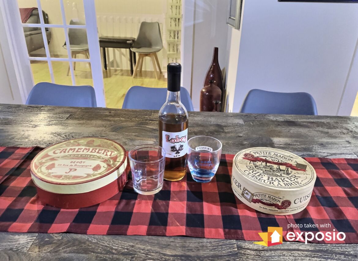 Een tafel met een rood-zwarte geruite tafelkleed, een fles Lobet, twee glaasjes en een blik Camembert.