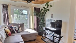 Un salon avec un canapé, un téléviseur posé sur un meuble, un ventilateur de plafond et une grande fenêtre avec des rideaux.