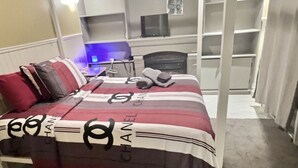 Une chambre d’hôtel avec un lit recouvert d’une courtepointe à rayures ornée du logo Chanel, un bureau avec un ordinateur, une chaise et un téléviseur posé sur une étagère.