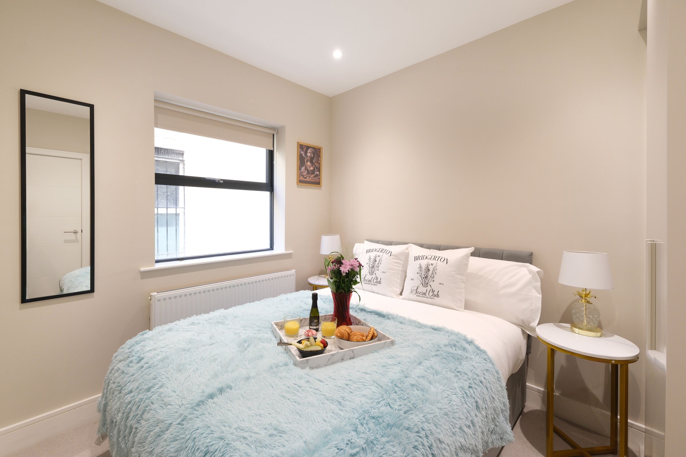 A bedroom with a large bed, a bedside table with a lamp, a window with blinds, a mirror, and a tray with breakfast items.