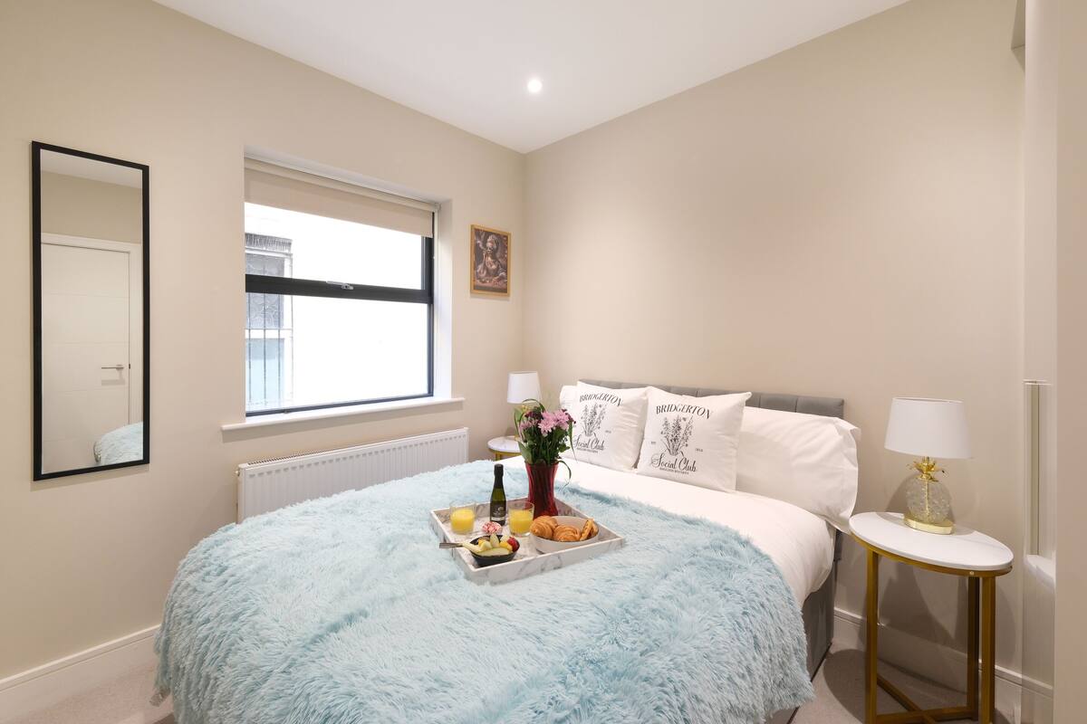 Un dormitorio con una cama grande, una mesita de noche con lámpara, una ventana con persianas, un espejo y una bandeja con alimentos para el desayuno.