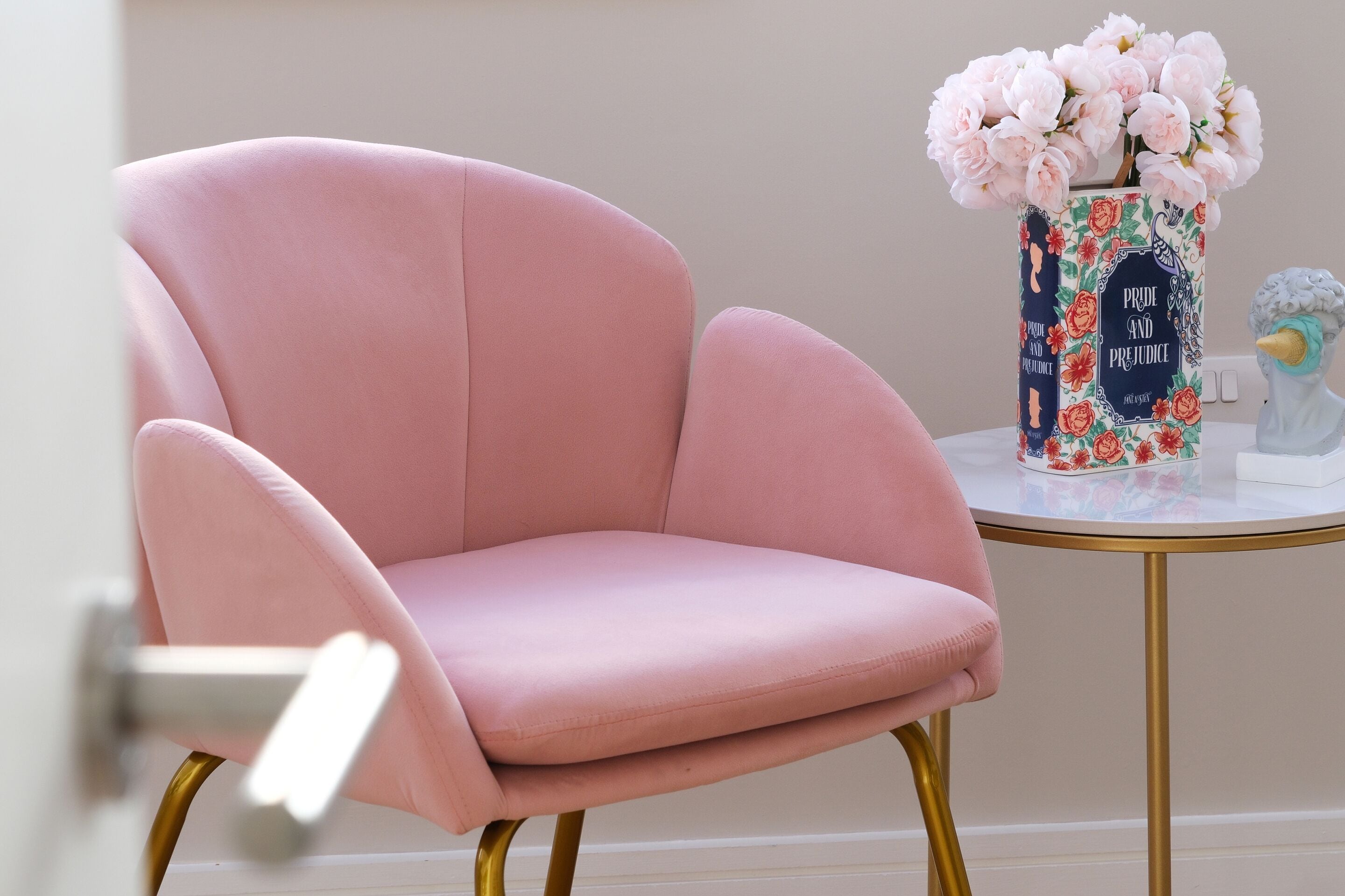 Un sillón rosa con estructura dorada, un jarrón con flores rosadas y un objeto decorativo sobre una mesita redonda.