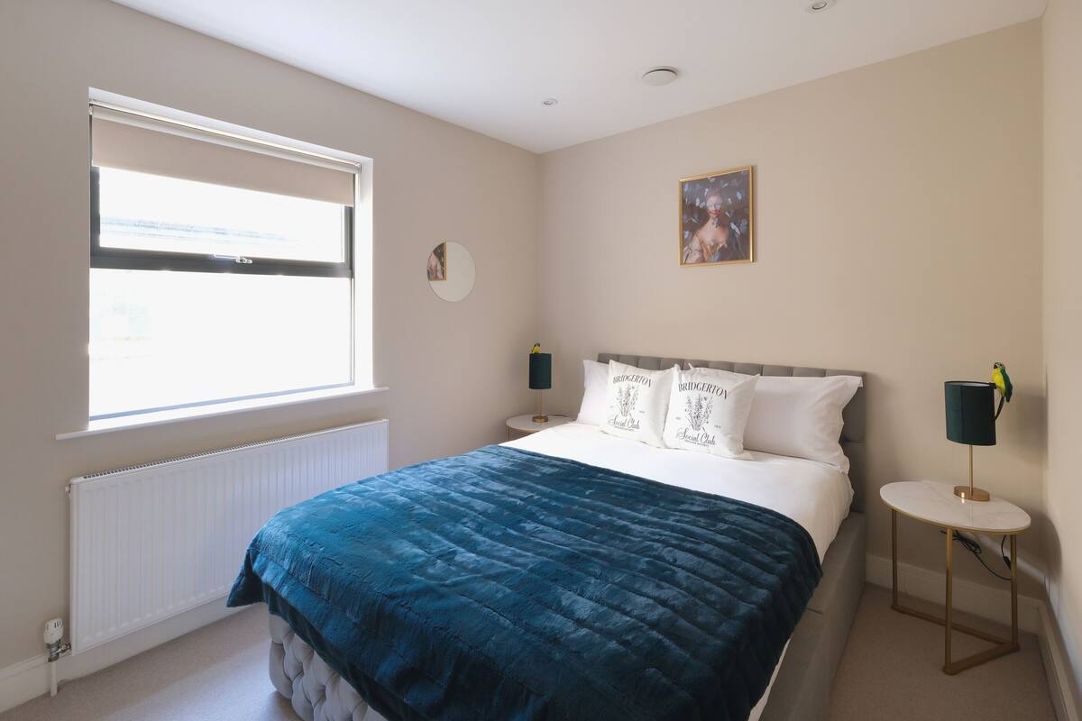 Un dormitorio con una cama grande, una ventana, un radiador, una mesita de noche con lámpara y un espejo montado en la pared.