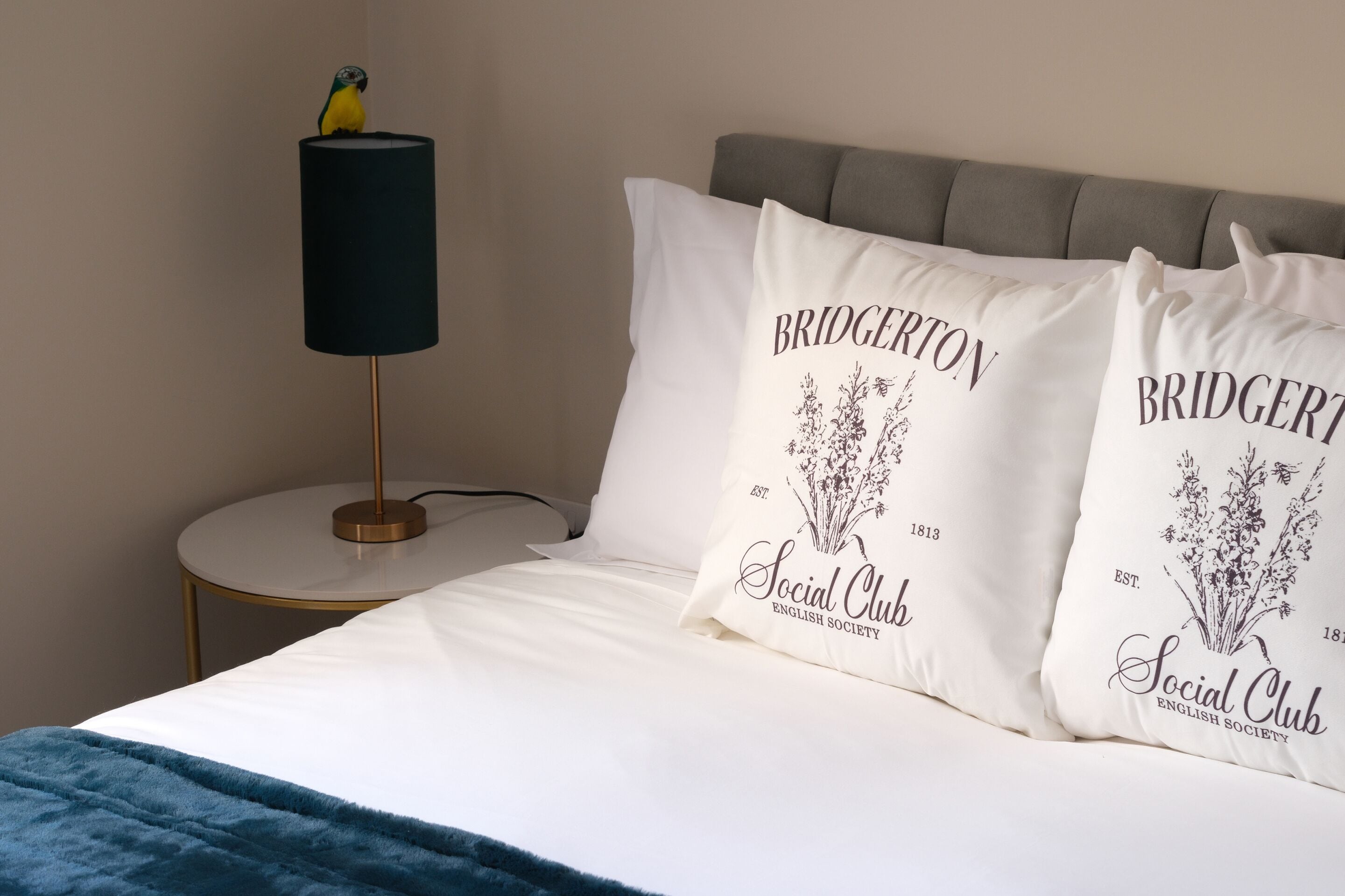 Una cama bien hecha con ropa blanca y dos almohadas con el logo de Bridgerton. Una lámpara con pantalla negra está sobre una mesita redonda junto a la cama.