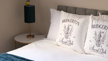 Una cama bien hecha con ropa blanca y dos almohadas con el logo de Bridgerton. Una lámpara con pantalla negra está sobre una mesita redonda junto a la cama.