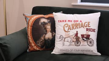 Un sofá verde con dos almohadones decorativos, uno con una ilustración de paseo en carruaje y texto, y el otro con un retrato antiguo.