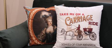 A green sofa with two decorative pillows, one with a carriage ride illustration and text, and the other with a vintage portrait.