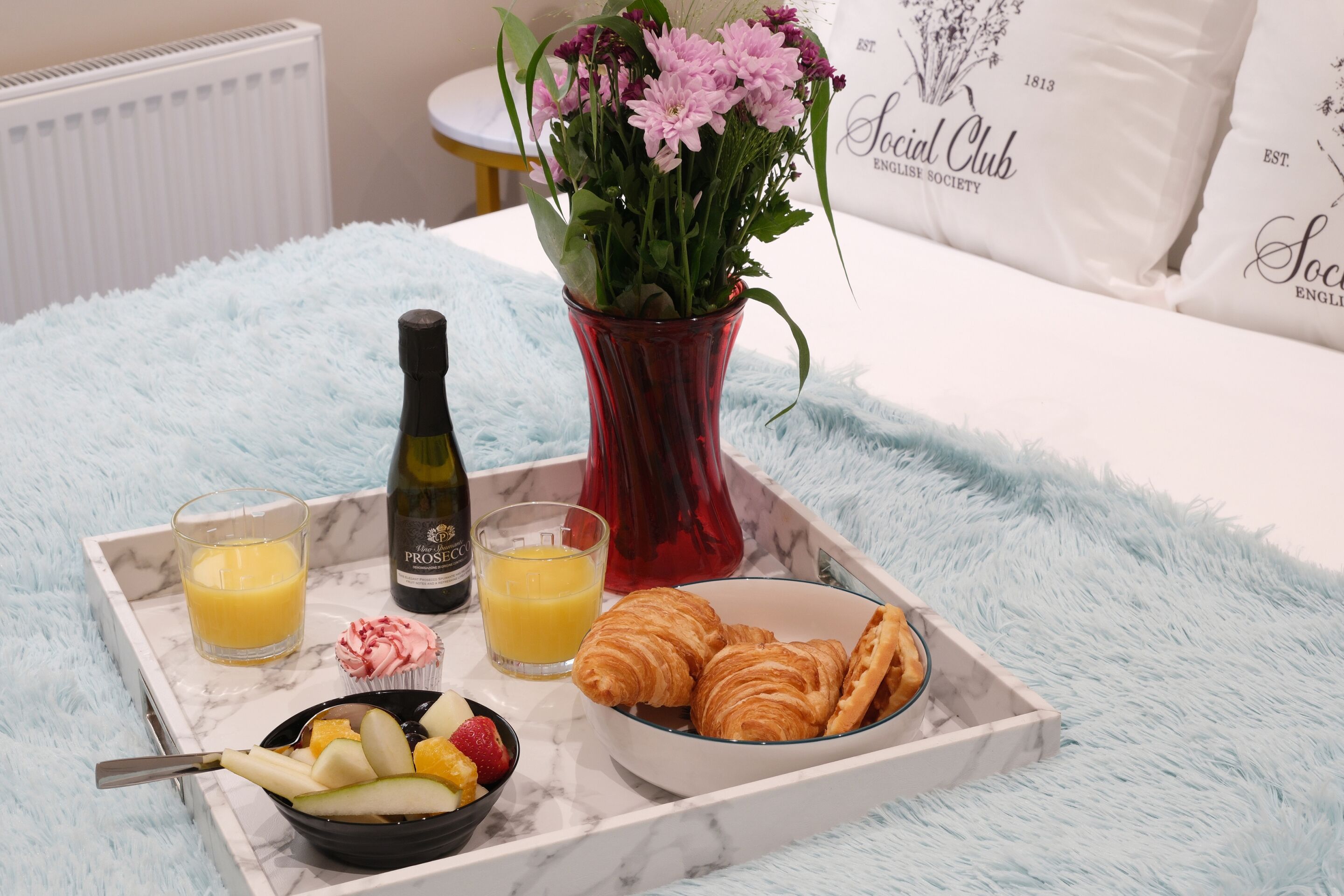 Un vassoio per la colazione con croissant, frutta, succo di frutta e una bottiglia di vino su una soffice coperta azzurra.