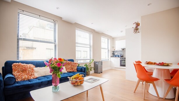 Ein modernes Wohnzimmer mit einem blauen Sofa, einem weißen Couchtisch und orangefarbenen Sesseln. Es gibt einen Essbereich mit einem weißen Tisch und orangefarbenen Stühlen.