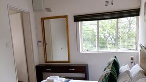 A neatly made bed with a blue and yellow bedspread, a large mirror with a gold frame, and a window with a view of trees. - SkyView, a spacious modern cottage. (Johannesburg)