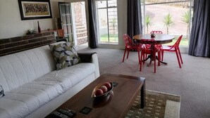 A living room with a sofa, a coffee table with fruit, a dining table with red chairs, and a view of the outdoors. - SkyView, a spacious modern cottage. (Johannesburg)