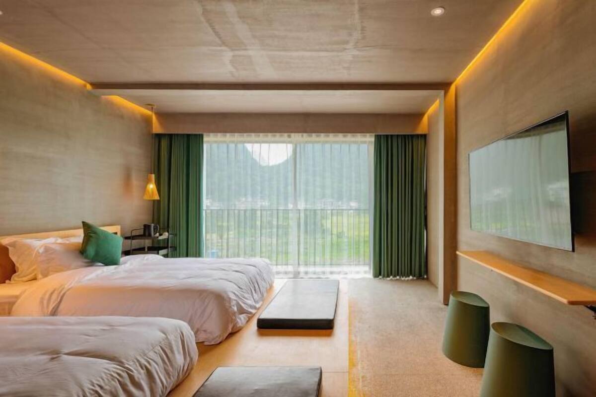 A modern hotel room with two beds, a large window with curtains, a flat-screen TV mounted on the wall, and a small seating area with stools.