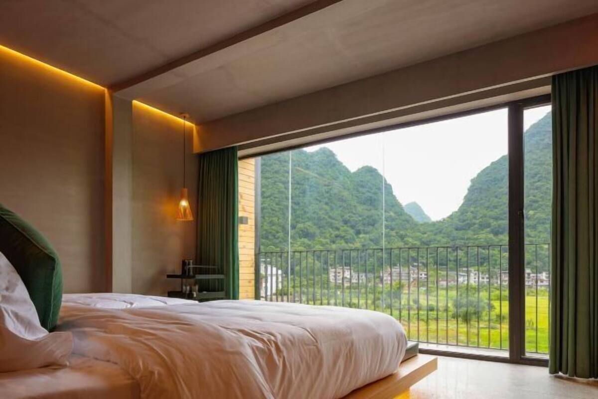 A bedroom with a large bed, a nightstand, a lamp, and a view of a mountainous landscape through a balcony.