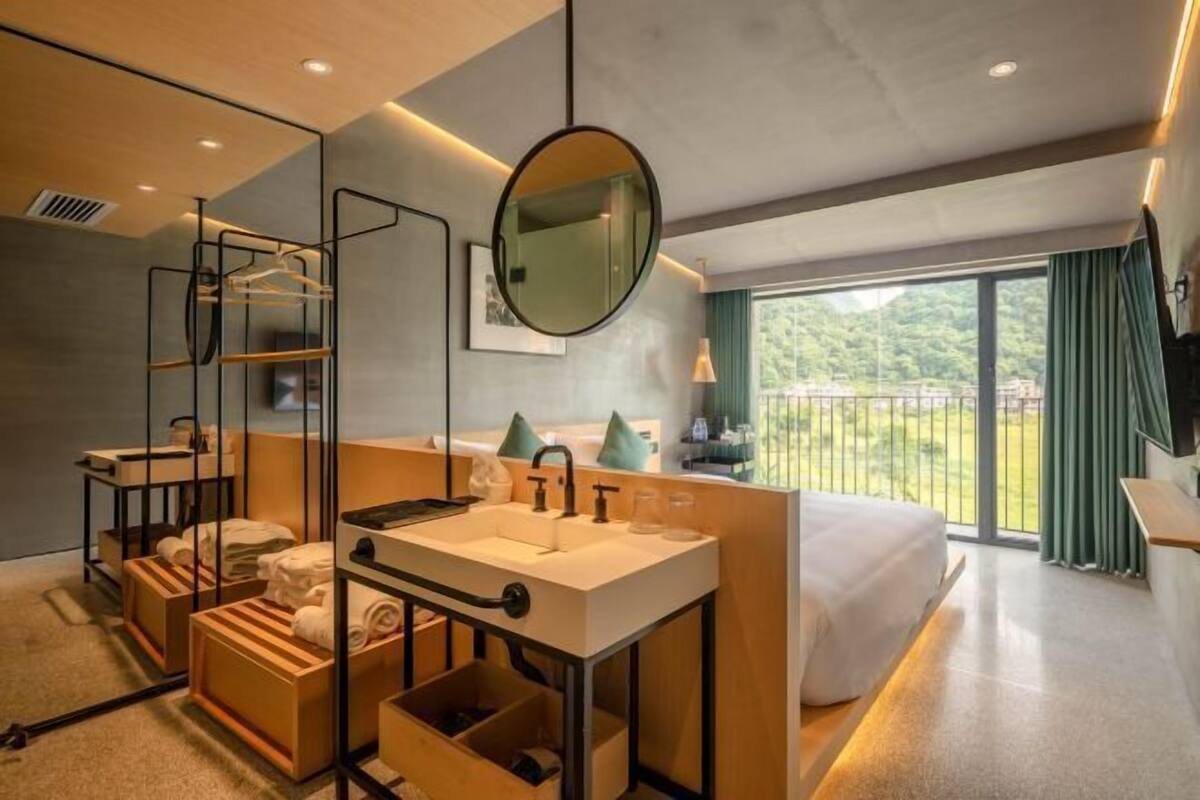 A modern hotel room with a large bed, a built-in vanity with a mirror, a window with a view of greenery, and a balcony.