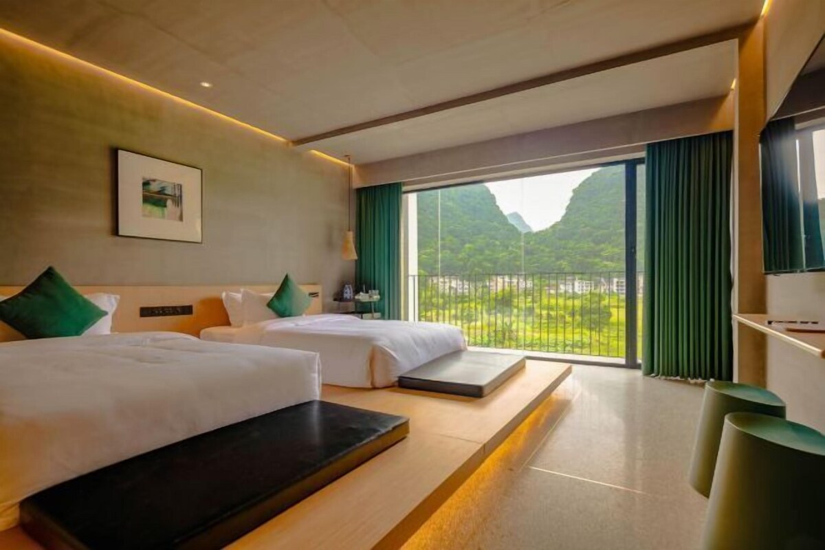 Ein Hotelzimmer mit zwei Betten, einem großen Fenster mit Blick auf Grünflächen und einem an der Wand befestigten Bild.