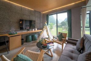 Ein modernes Wohnzimmer mit einer Couch, Sesseln, einem runden Tisch mit Früchten und Büchern, einem Teezelt, einem Flachbildfernseher und einem großen Fenster mit Blick auf Berge.
