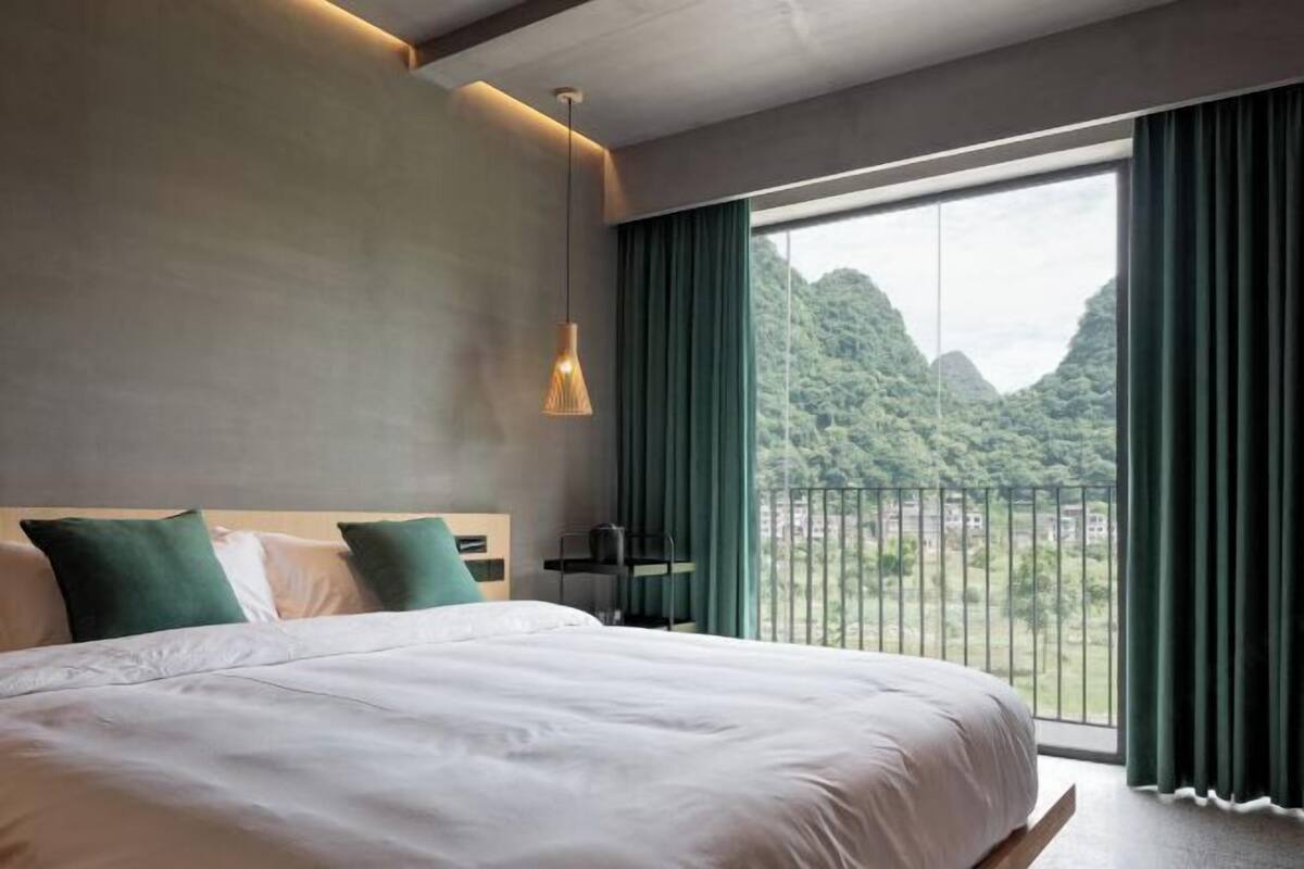 A modern hotel room with a large bed, a view of greenery, and a balcony.