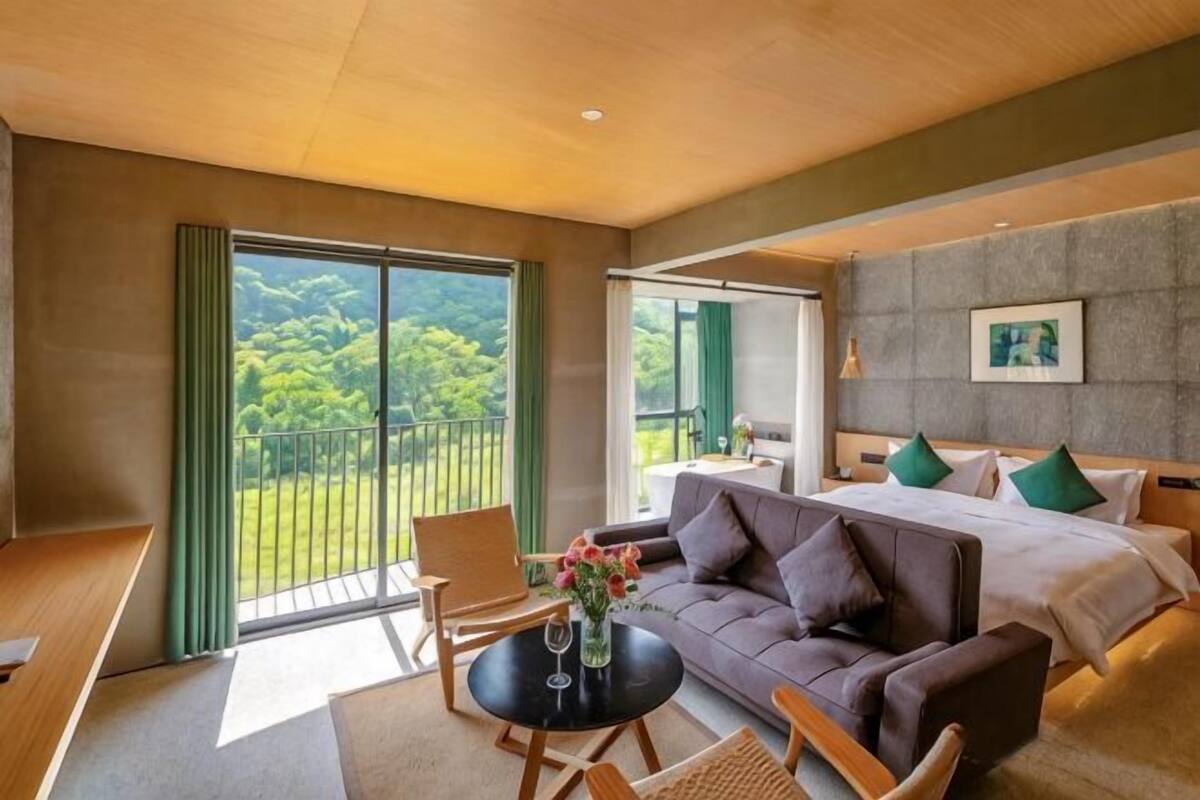 A hotel room with a balcony, a sofa, a round table with a vase, and a bed with pillows.
