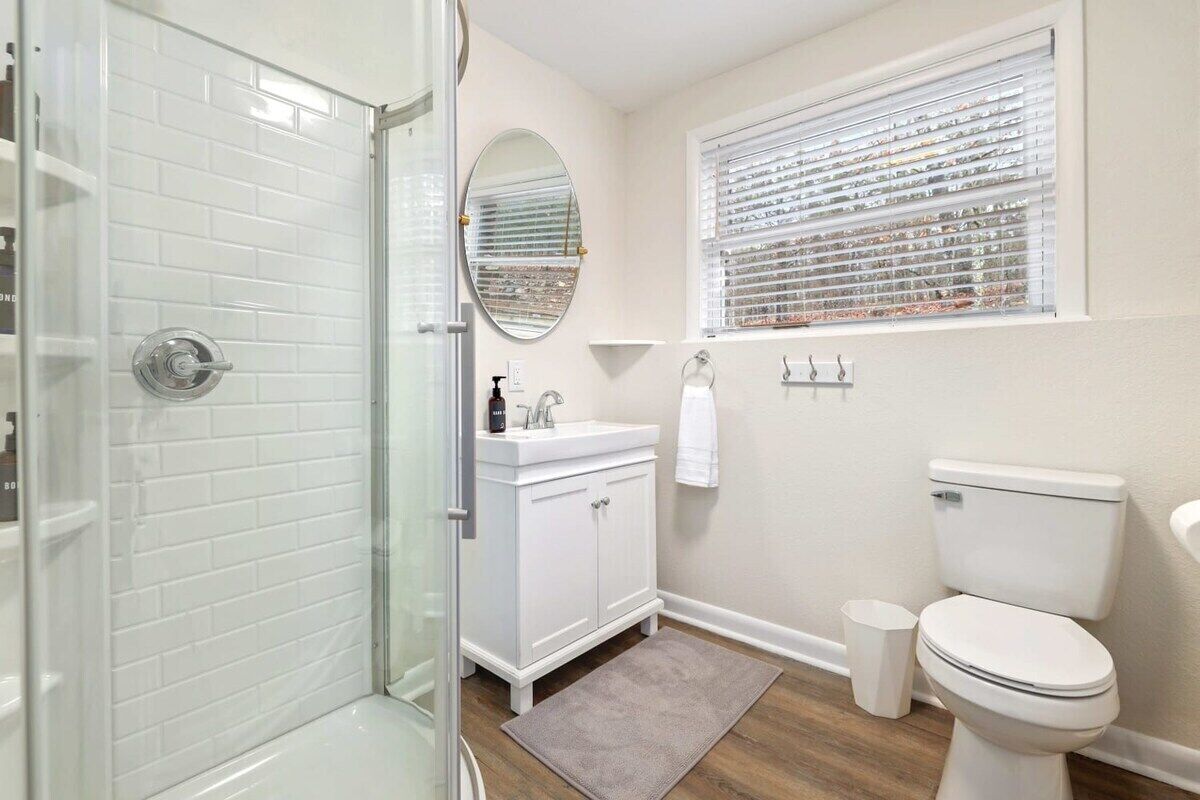 A bathroom with a glass shower enclosure, white tiled walls, a white sink with a mirror above it, a toilet, and a window with blinds.